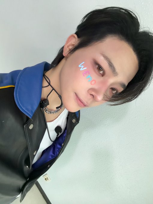 A selfie of a young man with dark hair wearing a black and blue jacket, with earphones around his neck and an AR effect displaying the text "weno" on his cheek.
