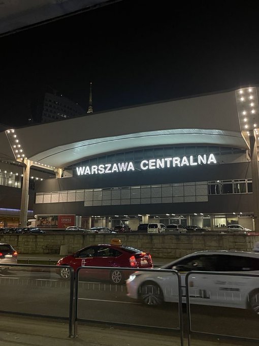 Nighttime view of the Warszawa Centralna train station entrance in Warsaw, Poland, with illuminated signage and cars passing in the foreground.