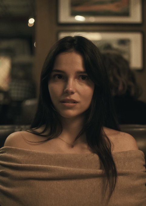 A young woman with long dark hair, wearing an off-shoulder taupe top and a delicate necklace, gazes directly at the camera with a serious, almost knowing expression in a dimly lit interior with framed artwork behind her—capturing the quiet triumph and restrained confidence of someone who made an early, lucrative investment.