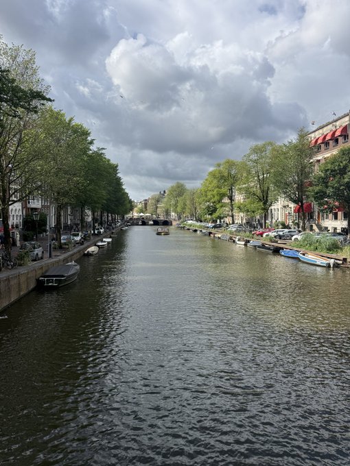 A scenic view of a canal lined with trees and buildings, with cars parked along the side and small boats on the water under a cloudy sky.