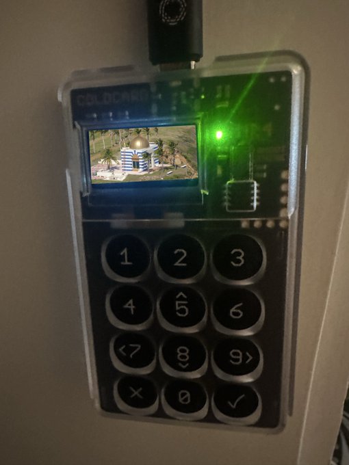 Close-up of a Coldcard Mk4 hardware wallet displaying a 'Burj Khalifa' Dubai screensaver image on its screen, with a green status LED lit, connected via a USB cable.