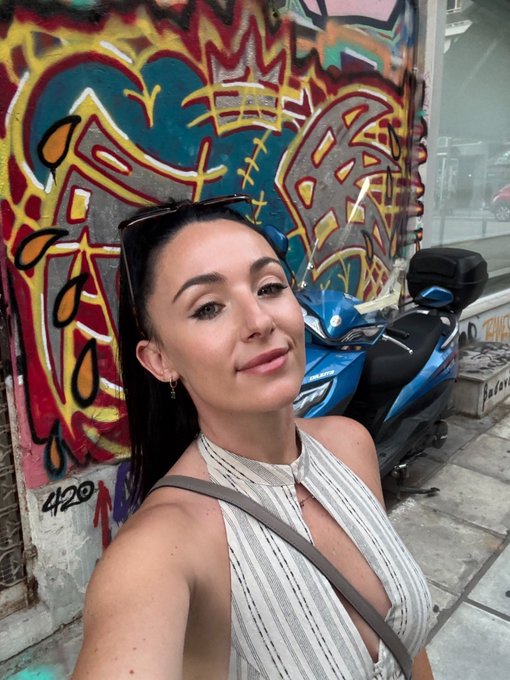 A woman with dark hair and sunglasses on her head takes a selfie in front of colorful graffiti and a blue scooter.