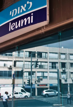 A street view shows a Leumi bank branch, identifiable by its blue and purple signage reading "לאומי" and "leumi" in both Hebrew and English. The image illustrates the Israeli banking giant that has announced it will offer cryptocurrency trading, including Bitcoin, Ethereum, and Solana, through its Leumi Trade app beginning in 2027.