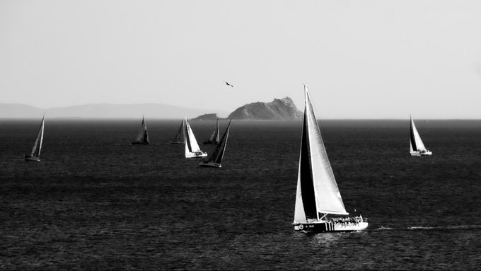 A black and white photograph of several sailboats on the open sea with a small island in the distance.