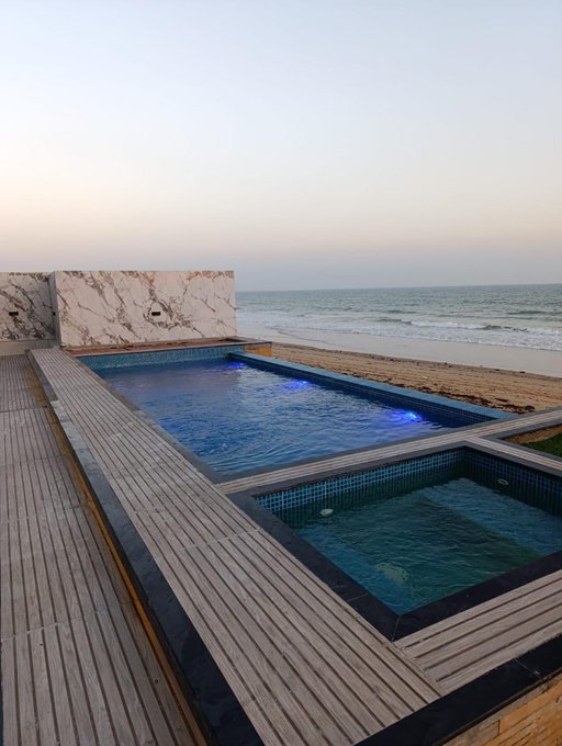 An infinity pool and spa on a wooden deck overlook the ocean at dusk, featuring a marble water feature wall and blue underwater lighting.