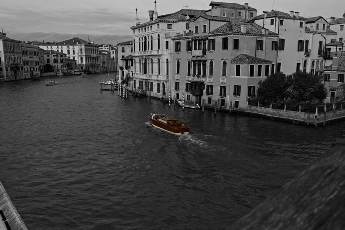 A black-and-white photograph of a Venetian canal lined with historic buildings, with a single boat highlighted in color as it travels down the waterway.
