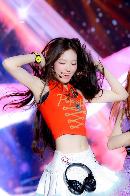 A K-pop idol performs on stage wearing a red halter crop top with 'Pepsi' written on it and a white skirt adorned with a large black bow.