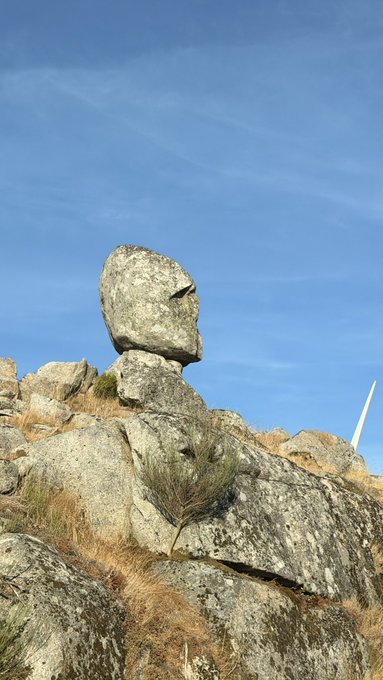 A balanced rock formation resembling a figure stands atop a rugged rocky outcrop against a bright blue sky, seemingly standing watch over the landscape. Fitting the "GM" (good morning) request, the natural stone sculpture appears ready to greet the day with its stoic presence.