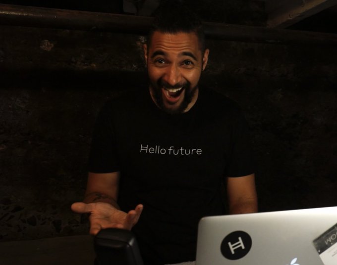A man with a beard and short hair excitedly laughs at a laptop screen while seated in a dimly lit room, wearing a black t-shirt with 'Hello future' printed in white text.