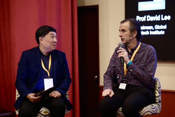 Two men sit on patterned chairs at what appears to be a panel discussion or interview, with one speaking into a microphone. A screen behind them identifies him as "Prof David Lee, Chairman, Global Fintech Institute," while the other man holds a tablet. The setting and professional attire with lanyards suggest a financial or fintech conference in Singapore, tying into the post's mention of Singapore-based discussions about cryptocurrency policy and private currencies.