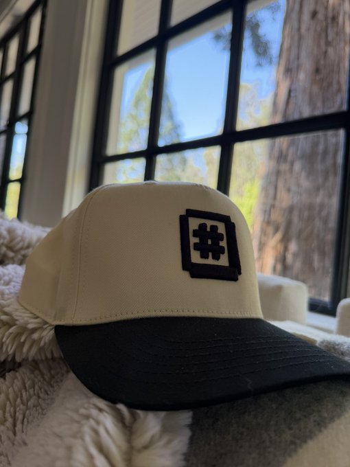 A tan and black baseball cap with a hashtag symbol patch sits on a fluffy blanket in front of a window with black gridded panes.