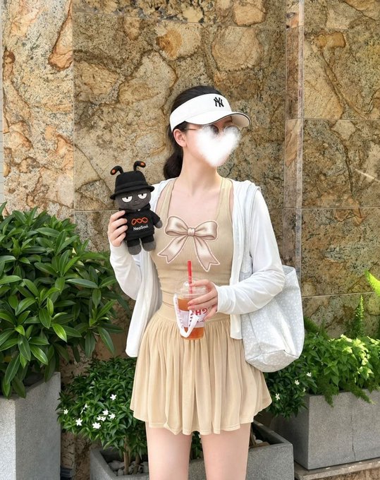 A person wearing a white NY visor, beige bow-detail dress, and white cardigan holds a black plush toy and an iced drink, standing in front of a marble-patterned wall with greenery.