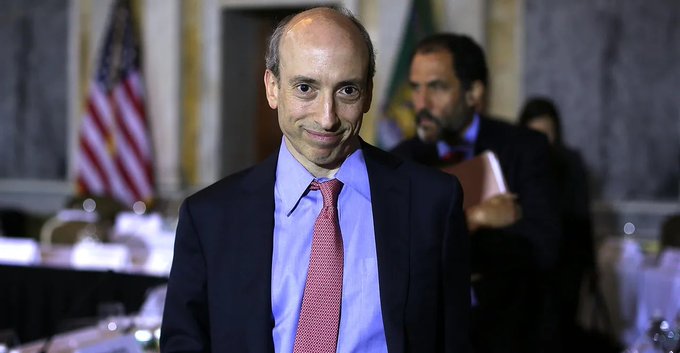 A photograph of a balding man in a dark blue suit, blue shirt, and red patterned tie smiling slightly, with an American flag visible in the background.