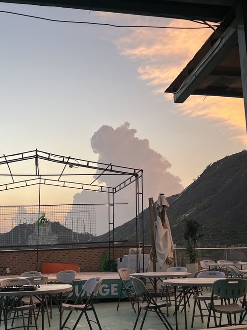 An outdoor patio with tables and chairs overlooks mountains and a dramatic cloud formation during a sunset sky.