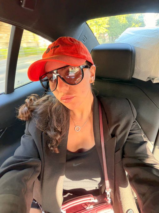 A selfie of a woman wearing a red 'op' baseball cap, dark sunglasses, a black blazer over a Nike tank top, and a pendant necklace, sitting in the back seat of a car with a red handbag on her lap.