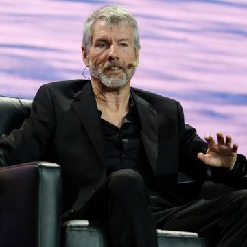 The image shows a man with gray hair and a beard, wearing a dark blazer over a black shirt, seated in a chair and gesturing with his hand as if speaking at a conference. The context suggests this is Michael Saylor, a well-known Bitcoin advocate, whose quote "Bitcoin is working today, So are we" aligns with the image of him likely delivering a speech or participating in a discussion about Bitcoin.