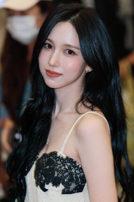 A young woman with long black wavy hair wearing a black and white dress with sequined details and red lipstick poses at what appears to be an event.
