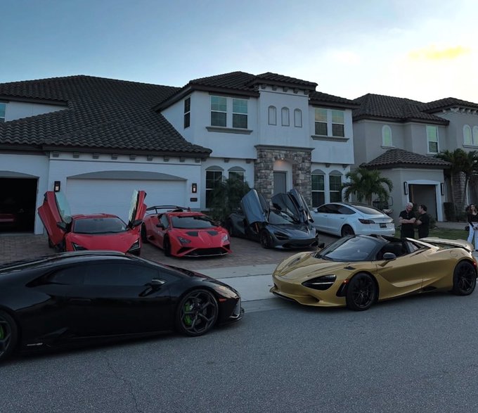 A collection of luxury sports cars parked in front of a large house, including a red Lamborghini Aventador with scissor doors open, a black McLaren, a gold McLaren 720S Spider, and a white Tesla.