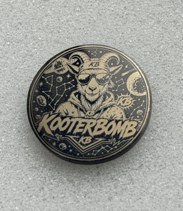 A round, metallic badge or coin rests on a gray textured surface, featuring an engraved design of a goat wearing sunglasses with the text 'KB' and 'KOOTERBOMB' surrounded by celestial motifs.
