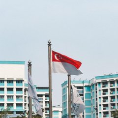 A Singaporean flag waves against a backdrop of white HDB public housing buildings under a pale blue sky, symbolizing Singapore as the regulatory authority shaping its stablecoin framework. The flag visually anchors the MAS's proposed policy shift toward allowing jointly issued stablecoins to qualify under its regulations.