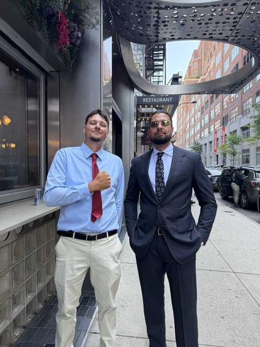 Two men in formal attire stand on a city sidewalk outside a restaurant; one wears a light blue shirt with a red tie, and the other wears a dark suit with sunglasses.