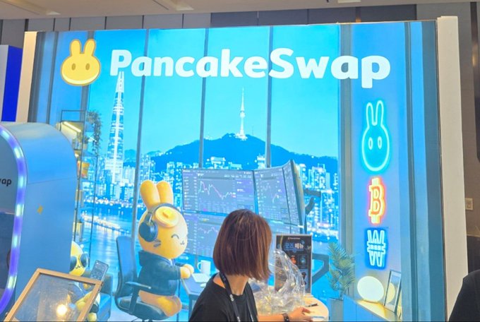 A blue-themed promotional booth for 'PancakeSwap' features a cartoon bunny mascot, a cityscape mural including Namsan Tower, and images of Bitcoin and other crypto symbols.