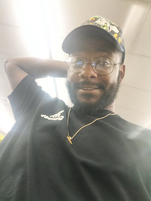 A selfie of a smiling man with a beard and glasses wearing a black t-shirt, gold chain, and a character-embroidered cap.
