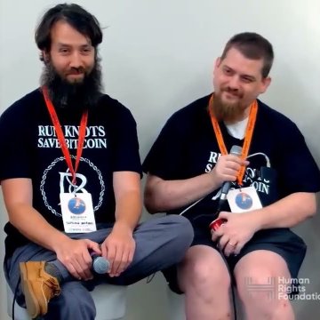 Two men sit side by side wearing dark blue shirts with 'RU KN0TS SAVE BITC01N' printed on them and lanyards around their necks, with a 'Human Rights Foundation' watermark visible.