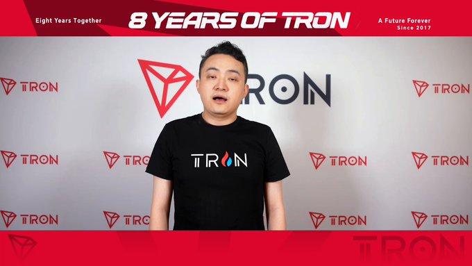 The image features a man wearing a black shirt with white text that says "TRX" written across his chest. He appears to be standing against a backdrop of red and white banners, which are likely part of an advertisement or promotional event related to TRON. The man is speaking into a microphone, suggesting he might be addressing the audience at this event.