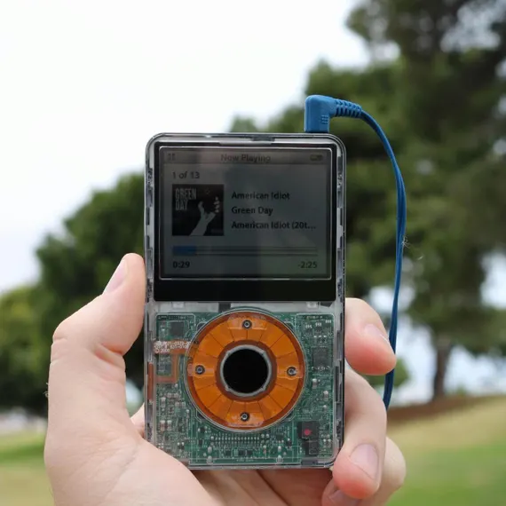 A close-up of a transparent portable media player with exposed circuit boards, displaying a 'Now Playing' screen for 'Green Day - American Idiot' in a person's hand.