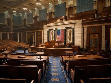 Interior view of the U.S. House of Representatives chamber, showing the speaker's rostrum, American flag, and tiered seating desks.