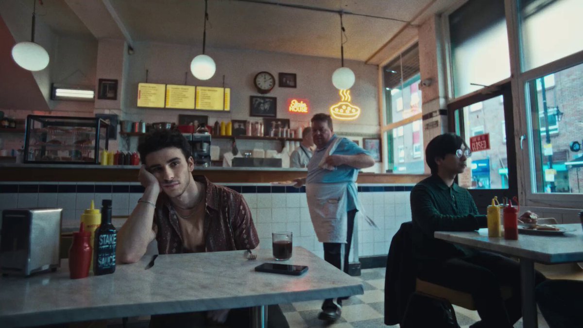 The image features a group of people sitting around dining tables, with one person sitting alone at a table looking bored while others are engaged in conversation or eating. The scene takes place inside a restaurant, where there are multiple tables and chairs arranged throughout the space. A clock is visible on the wall above the diners' heads.

The post includes an image of a man wearing a blue shirt with the words "Pizza" written on it, suggesting that they might be at a pizza parlor or a restaurant serving pizza. The caption reads: "All at stake this summer Hazard coaching the legends in Stake's new summer ad is pure genius drop your bold winner prediction below".