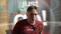 A man in a red T-shirt speaks and gestures in an interview setting; the word "just" is visible in the background and on his shirt.