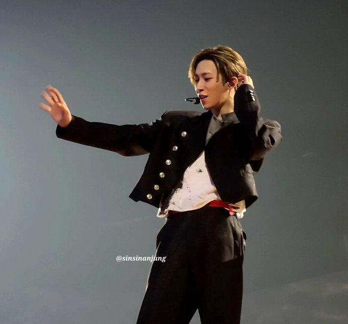 A K-pop performer in a black military-style cropped jacket with gold buttons and red sash, performing on stage with arms outstretched, with watermark '@sinsinanjung'.