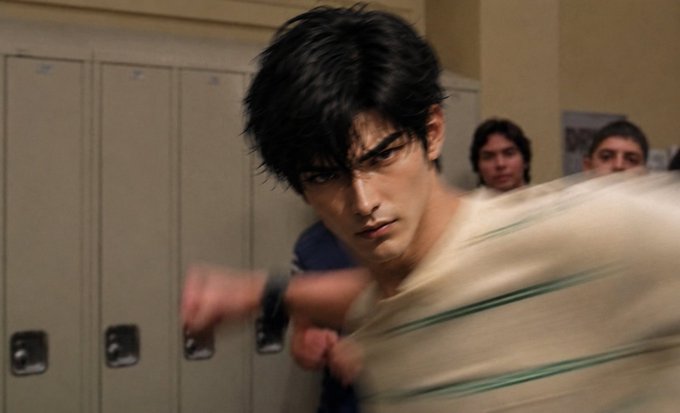 A scene showing a young man with dark hair in an intense, aggressive pose with motion blur, appearing to throw a punch in what looks like a school hallway with lockers and other people in the background.