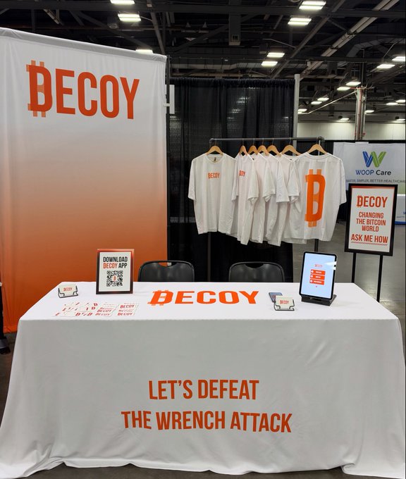 A trade show booth for 'DECOY' displays t-shirts with the Bitcoin logo, a table with a 'LET'S DEFEAT THE WRENCH ATTACK' tablecloth, and signage reading 'CHANGING THE BITCOIN WORLD, ASK ME HOW.'