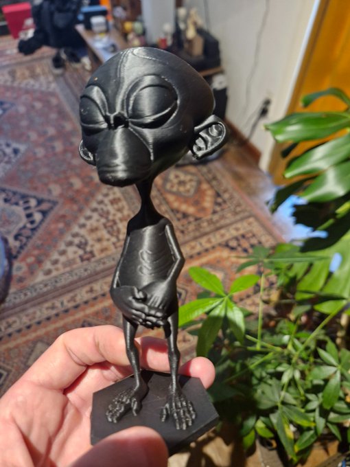 A hand holds a black figurine of a monkey-like alien with large eyes and a slender body, photographed indoors near a patterned rug and green houseplant.