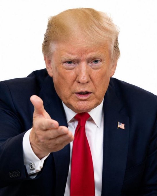 A close-up photo of Donald Trump wearing a dark suit, white shirt, and red tie, pointing forward against a white background.