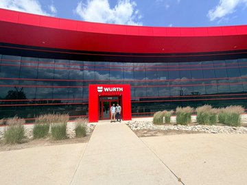 A photograph of a modern building with a red curved glass facade and a 'WURTH' sign above the entrance, with people walking toward the door.