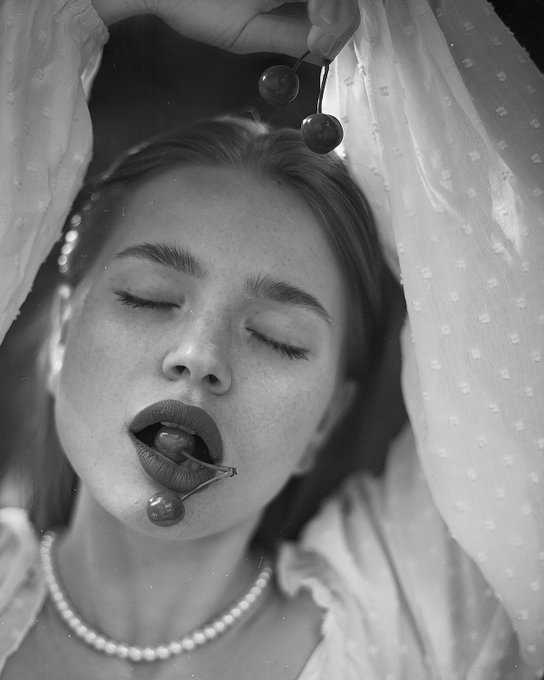 A black and white close-up portrait of a woman with closed eyes, freckles, and cherries held near her mouth and hair.