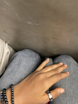 A close-up photo shows a person's hand resting on their gray trousers, with a black beaded bracelet and a silver ring visible, set against a concrete floor. The caption suggests they are en route to the @NFT__NG event, hinting at anticipation while wondering if others will attend.