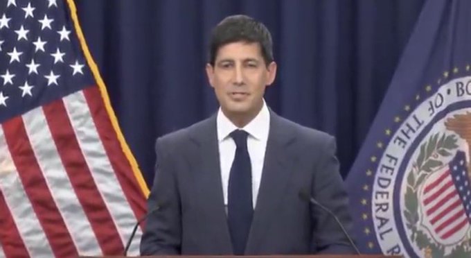 A man in a dark suit stands at a podium with the American flag and the seal of the Federal Reserve Board visible behind him, suggesting an official Federal Reserve press briefing. The post about July CPI coming in at 2.7% below the 2.8% forecast ties into this Federal Reserve context, as the official pictured appears to be delivering commentary relevant to inflation data and potential interest rate decisions.
