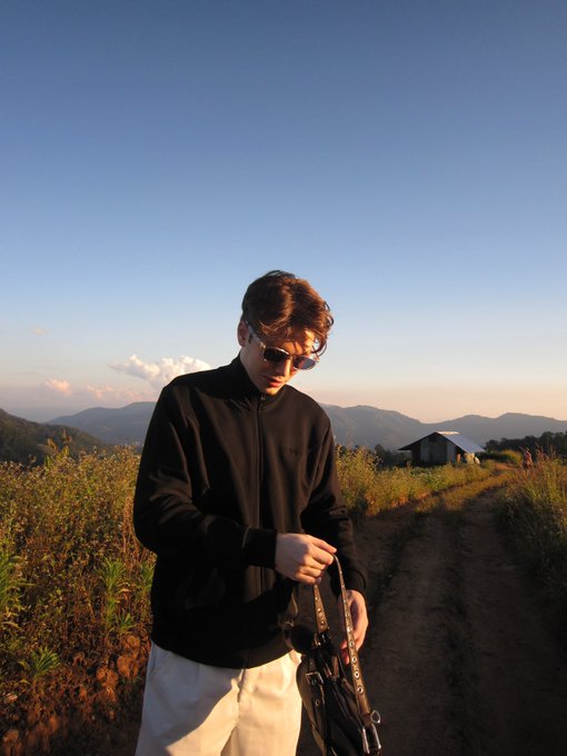 A young man wearing sunglasses and a black jacket stands on a dirt path with mountains in the background, looking down while adjusting a bag strap. The image pairs with an introductory post where he's identified as "Lorenzo2k," a Thailand-based crypto enthusiast sharing his background and interests.