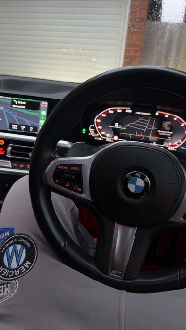A BMW M Sport steering wheel with the iconic blue and white roundel is shown from the driver's seat, featuring an M badge on the lower spoke, an M-specific digital instrument cluster, and a "Welcome" message on the central infotainment screen, with a "Neil Murley Automotive" watermark visible on the driver's clothing — humorously captioned "GM bros," playing on the contrast between BMW's M performance division and a GM enthusiast.