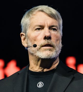 The image features a man wearing a black suit with a microphone in his mouth, standing against a dark background. The caption of the image reads "MICHAEL SAYLOR: Bitcoin will surge to $180,000 and crash to $140,000, people will be freaking out about it again." This statement is related to the content of the post by Michael Saylors, who has predicted that Bitcoin's value will increase rapidly and then crash. The man in the image appears to be discussing this topic with someone else or presenting his views on the matter.