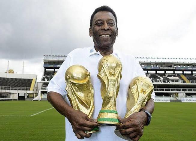 A smiling man, who appears to be Pelé, holds three FIFA World Cup trophies while standing on a football pitch in front of a stadium.