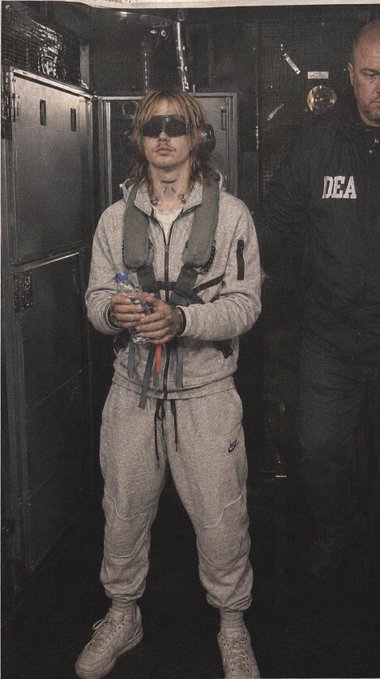 A person wearing dark sunglasses, a gray tracksuit, and a tactical vest stands holding a water bottle, accompanied by an agent wearing a black 'DEA' jacket in a dimly lit industrial setting.