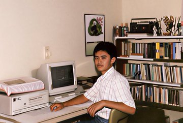 A vintage photo of a young man with dark hair wearing a white striped shirt sitting at a desk with an old CRT monitor, printer, and a bookshelf filled with books in the background.