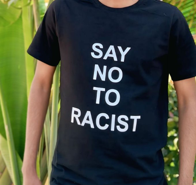 A person wearing a black t-shirt with white text that reads 'SAY NO TO RACIST', standing outdoors near green plants.