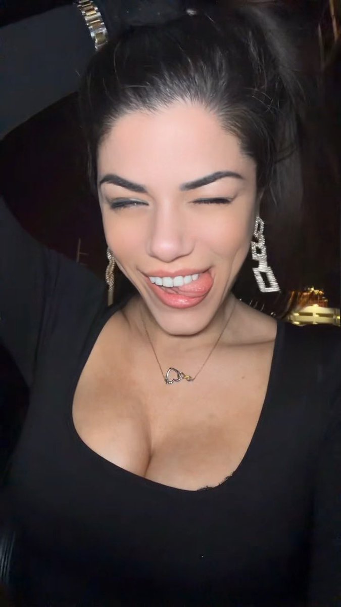 A smiling woman with dark hair pulled back, wearing a low-cut black top and large dangling earrings, sticks her tongue out playfully at the camera.